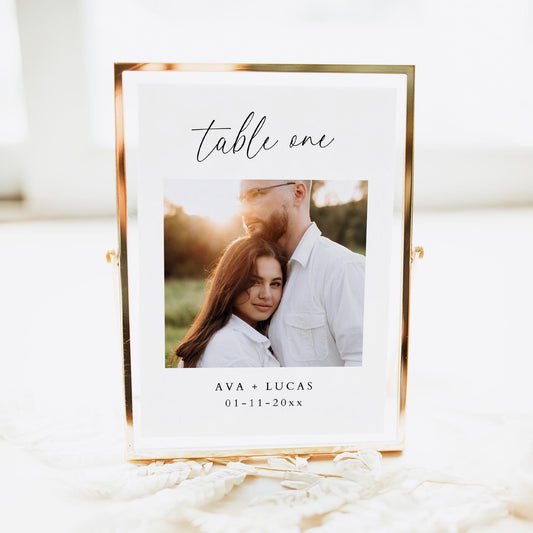 Table number card in a frame featuring the text &#39;table one&#39; in script font, a photo of the couple, their names and the wedding date